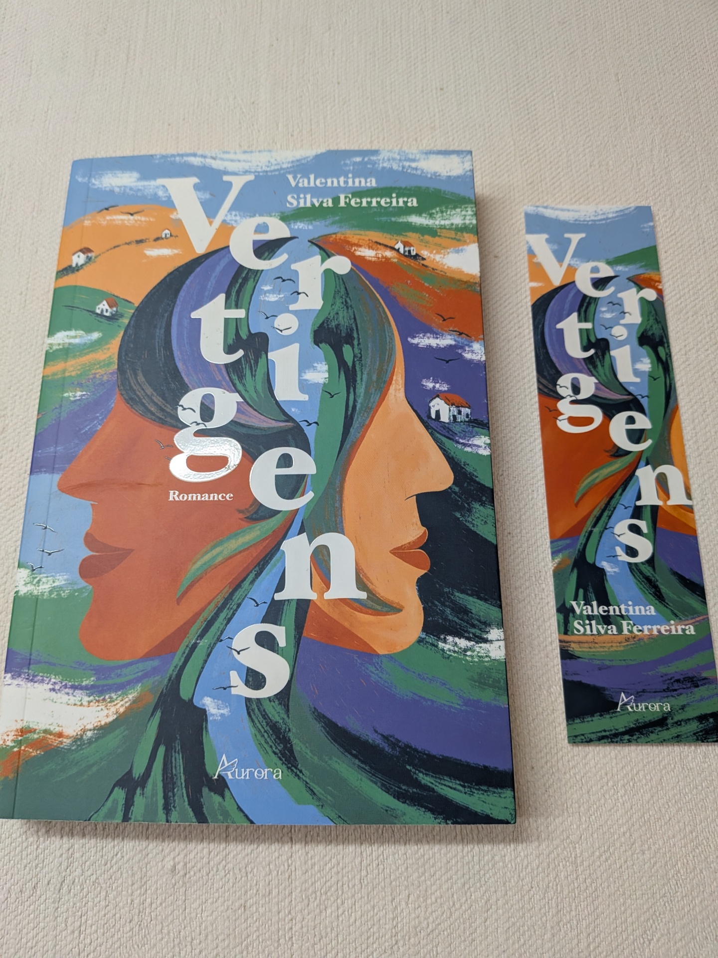 Valentina Silva Ferreira - Vertigens Livro e marcador de livro com capa colorida e arte abstrata de rostos na paisagem