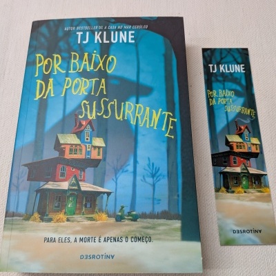 Livro 'Por Baixo Da Porta Sussurrante' e marcador de livro com capa em tons azuis e amarelos com ilustração de casa e mota