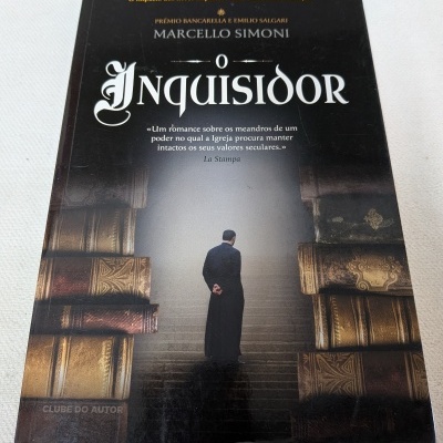 Livro O Inquisidor de Marcello Simoni com capa preta, pessoa de costas vestindo hábito, e pilhas de livros antigos dourados.