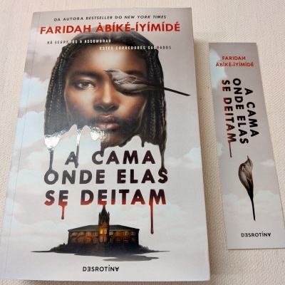 Livro 'A Cama Onde Elas Se Deitam' com rosto feminino e pássaro na capa, ao lado marcador de páginas.