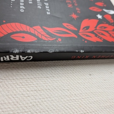 Livro com capa preta e detalhes vermelhos e brancos, padrão de folhas e estrelas, texto 'CARRIE' e 'STEPHEN KING', desgaste visível na capa
