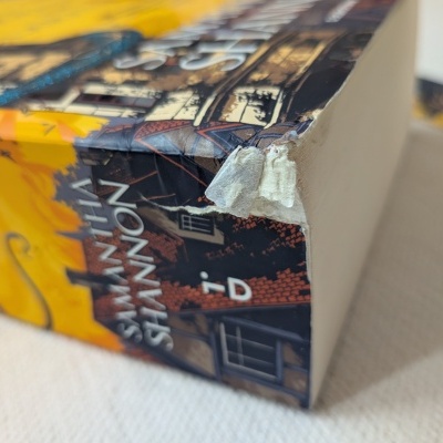 Caixa de livro em cartão com canto danificado e ilustração amarela e azul.