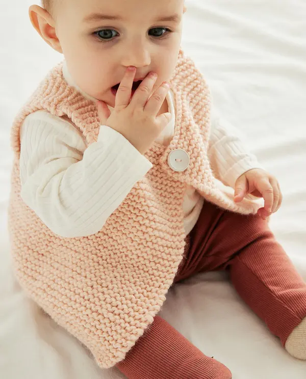 Bebé sentado com colete rosa de malha, camiseta branca e calças ferrugem