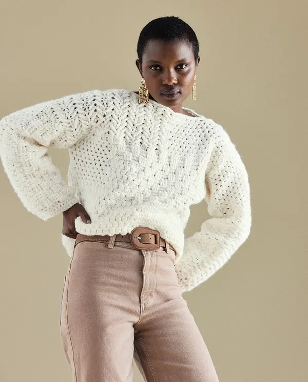 Camisola branca de malha com padrão aberto e calças rosadas com cinto castanho em fundo neutro