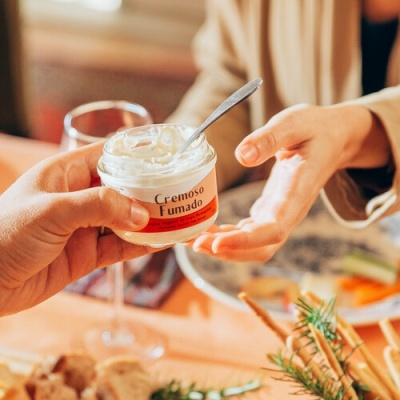 Frasco de creme 'Cremoso Fumado' sendo servido à mesa com comida e bebida ao fundo