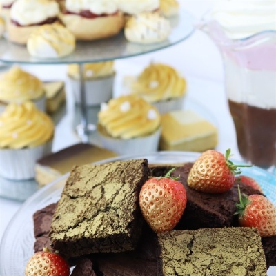 Sobremesas variadas incluindo brownies com pó dourado e morangos, mini cupcakes amarelos, e jarra com líquido marrom e rosado.