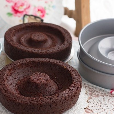 Bolos de chocolate em forma de anel sobre suporte decorado e toalha de renda branca ao lado de formas de alumínio