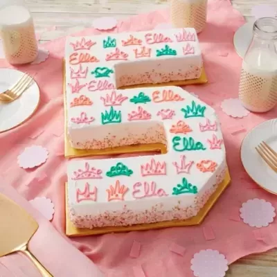 Bolo número 5 decorado com glacê branco e colorido com escrita e desenhos, sobre mesa com toalha rosa e utensílios de mesa.