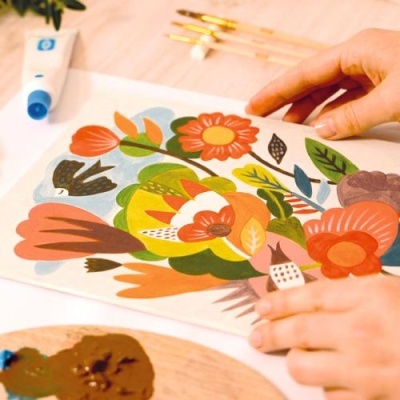 Folha com pintura colorida de flores sendo segurada por mãos, pincéis e tubo de tinta ao fundo