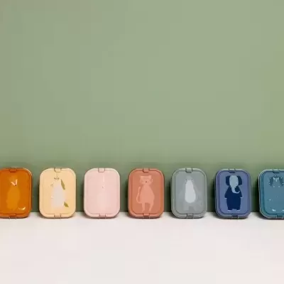 Oito caixas metálicas coloridas com recortes de animais na tampa, alinhadas sobre superfície branca e fundo verde.