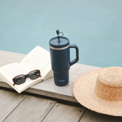 Copo térmico azul com pega e canudo, chapéu de palha, livro aberto e óculos de sol junto à piscina