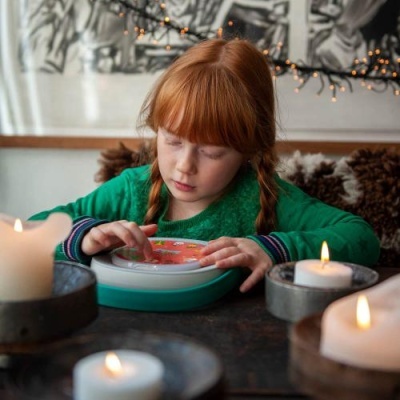 Criança com camisola verde a usar dispositivo redondo colorido numa mesa com velas acesas