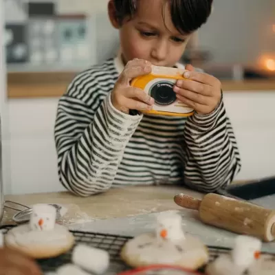Menino segurando uma câmara fotográfica digital amarela e branca numa cozinha com bolachas decoradas e utensílios de cozinha