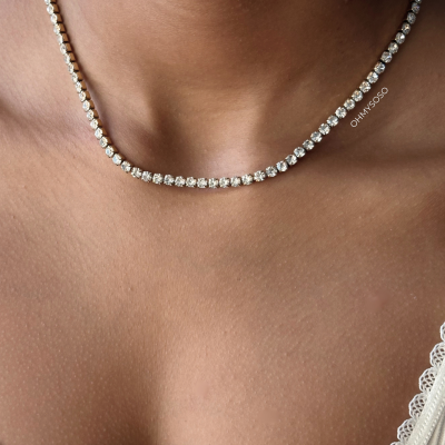 Colar tennis dourado com pedras transparentes redondas em pescoço de pessoa vestindo branco canelado com bordado.