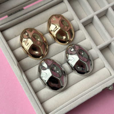 Botões metálicos ovais dourados e prateados numa caixa cinzenta de veludo sobre fundo rosa.