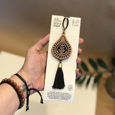 Colar em madeira formato gota com pendente tassel preto, apresentado em placa branca com texto em português