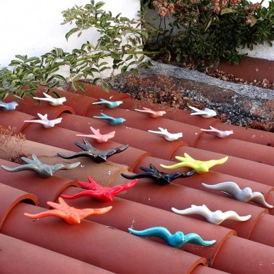 Pequenas esculturas cerâmicas de pássaros coloridos no telhado de telhas vermelhas com vegetação ao fundo