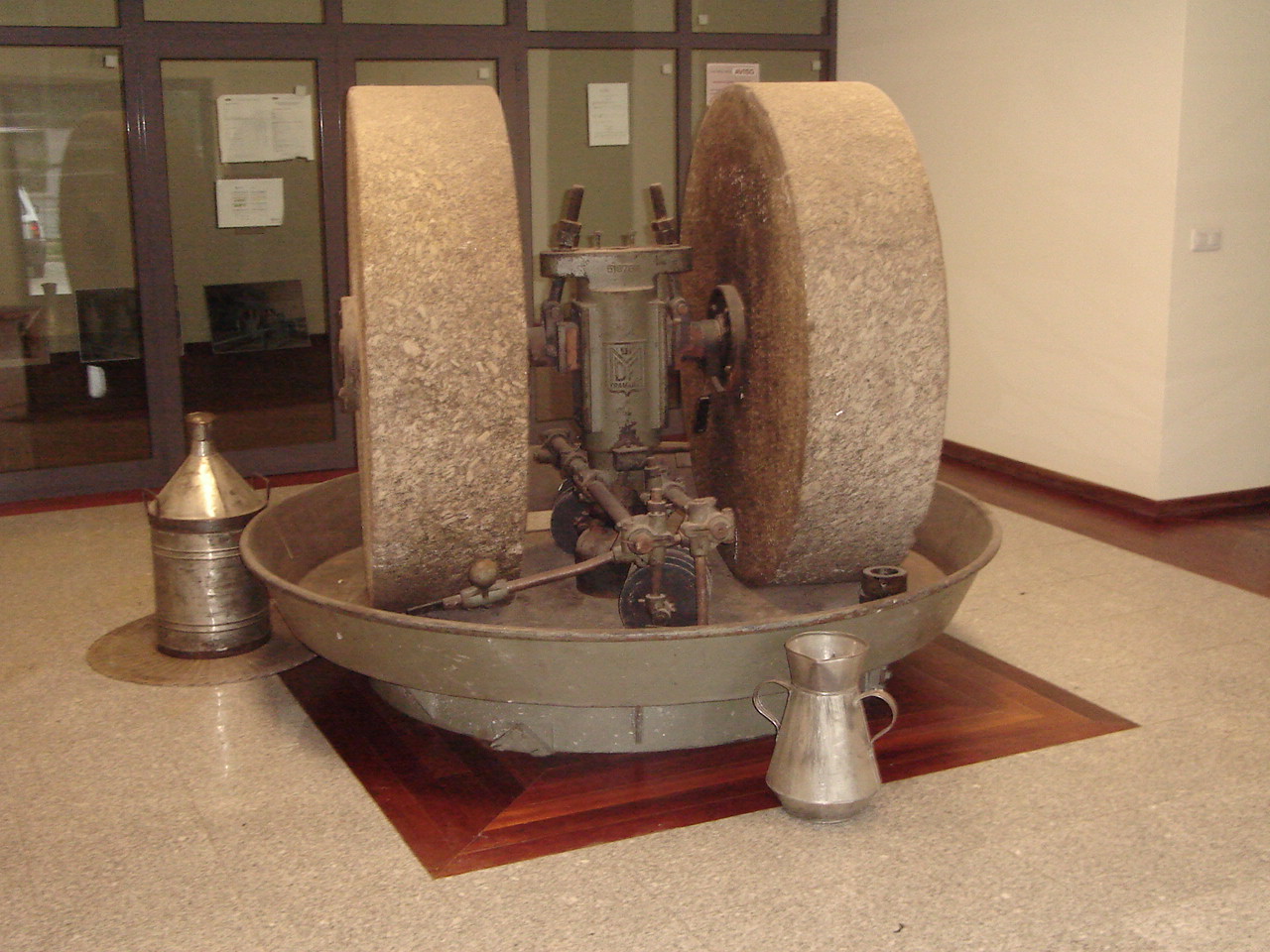 Máquina antiga com rodas de pedra e recipientes metálicos em chão de madeira e mármore