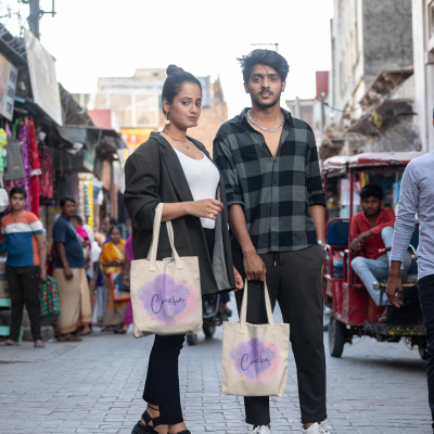 Dois jovens segurando sacolas de tecido bege com padrão roxo e azul e texto 'Creche' numa rua movimentada.