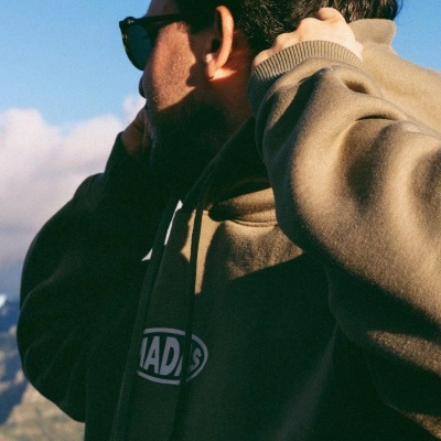 Pessoa vestindo sweatshirt verde-oliva com capuz e texto branco MADS, ao ar livre com céu azul e montanhas.