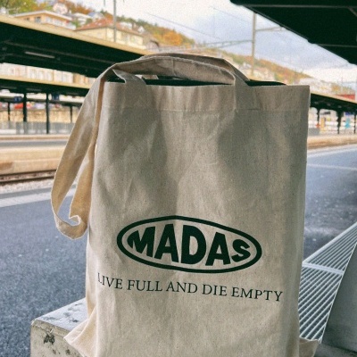 Saco tote bag de algodão cru com estampado verde 'MADAS' e frase, numa estação de comboios