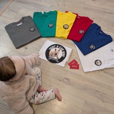 Sete t-shirts coloridas com imagem de cão e texto RENT A CACHORRO, bebé ao lado sentado no chão de madeira