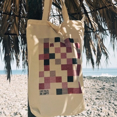 Saco de tecido bege com padrão geométrico e texto pendurado em estrutura de madeira numa praia rochosa