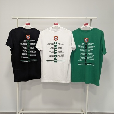 Três t-shirts preta, branca e verde com brasão de Portugal e lista de nomes nas costas penduradas numa estrutura branca