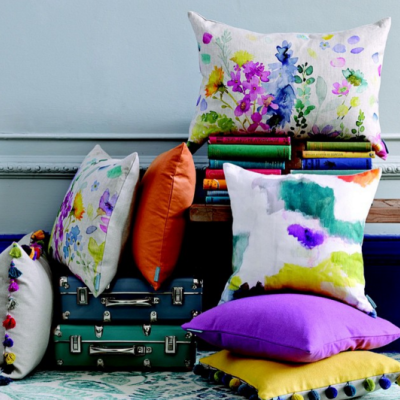 Almofadas coloridas decorativas com padrões florais e manchas, empilhadas sobre malas e livros em fundo cinza claro.