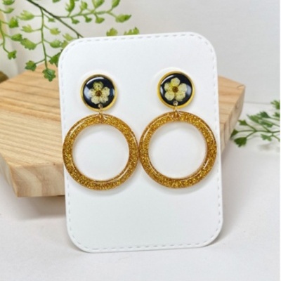 Brincos dourados com flores secas em fundo preto, pendurados em cartão branco