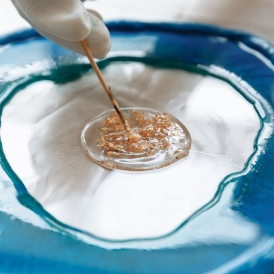 Mistura de resina epóxi transparente com partículas douradas num molde azul e branco, mãos com luva branca a mexer com palito de madeira