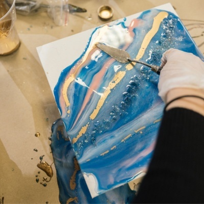 Tela abstrata com resina azul, rosa, branca e dourada e textura em relevo, com mãos usando luva a trabalhar com espátula.