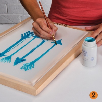 Pessoa pintando setas azuis em tecido branco numa moldura de madeira com pincel e frasco de tinta