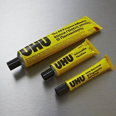 Três tubos amarelos de cola UHU com texto em várias línguas sobre superfície metálica.