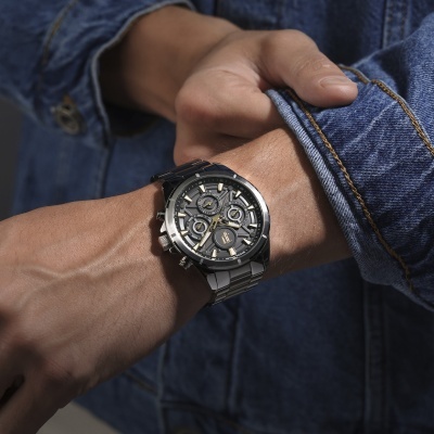 Relógio de pulso metálico com mostrador preto e detalhes dourados no pulso de uma pessoa com jaqueta de ganga azul