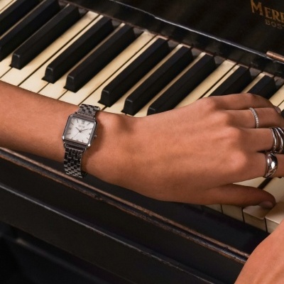 Relógio de pulso quadrado com bracelete de metal prateado numa mão sobre um piano preto.