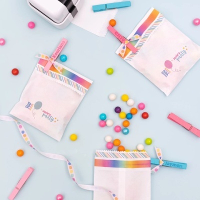 Três sacos de tecido branco com faixa colorida e texto, pregadores de madeira coloridos e doces coloridos em fundo azul claro.