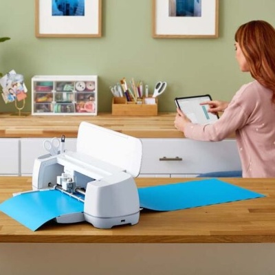 Impressora branca a imprimir folhas azuis numa bancada de madeira clara com mulher a usar tablet ao fundo