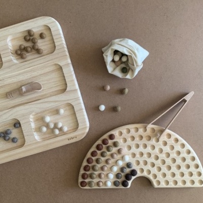 Jogo de madeira com bolas coloridas, tabuleiros e pinça, sobre superfície castanha.