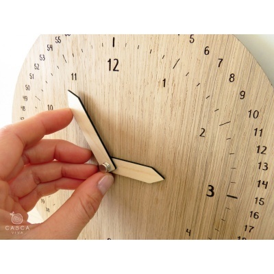 Relógio de parede em madeira clara com numeração preta e ponteiros em madeira sendo ajustados por uma mão humana