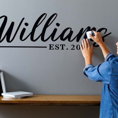 Mulher aplicando adesivo de parede preto com palavra 'Williams EST. 20' em parede cinza acima de prateleira de madeira com livros e objeto decorativo branco