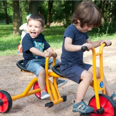 crianças a brincar num triciclo amarelo e vermelho num parque exterior