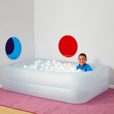 Piscina insuflável branca com bolas brancas e menino vestido de azul dentro, em sala com tapete cor-de-rosa e espelhos coloridos na parede