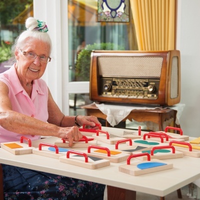 Idosa a jogar jogo de tabuleiro de madeira com peças coloridas numa sala com rádio antigo