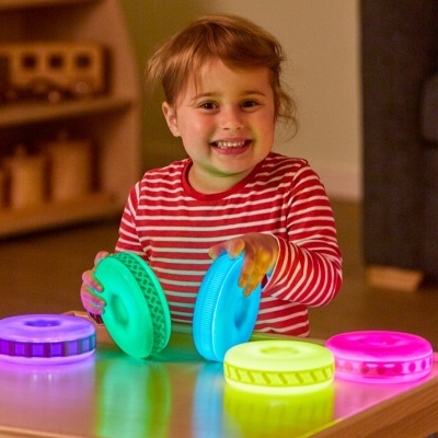 Criança com camisa riscada brincando com discos luminosos coloridos numa mesa.