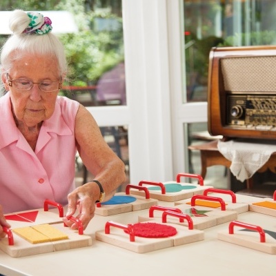 Mulher idosa usando um jogo de encaixe em madeira com alças vermelhas, sentada à mesa em ambiente interno.