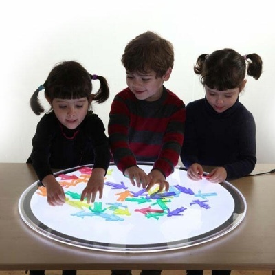 Três crianças a brincar com figuras coloridas numa mesa redonda iluminada