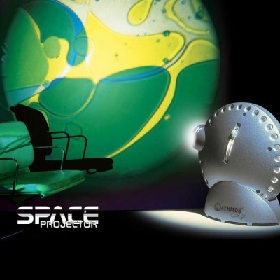 Projetor metálico prateado a projetar padrão colorido numa parede com cadeiras verdes ao lado