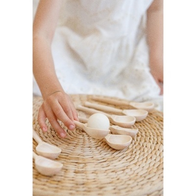 Colheres de madeira claras dispostas em círculo sobre superfície de palha com criança a tocar uma colher com bola branca