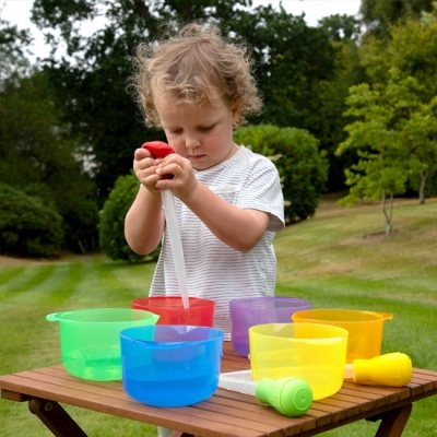 Criança pequena brincando com recipientes plásticos coloridos em mesa de madeira ao ar livre